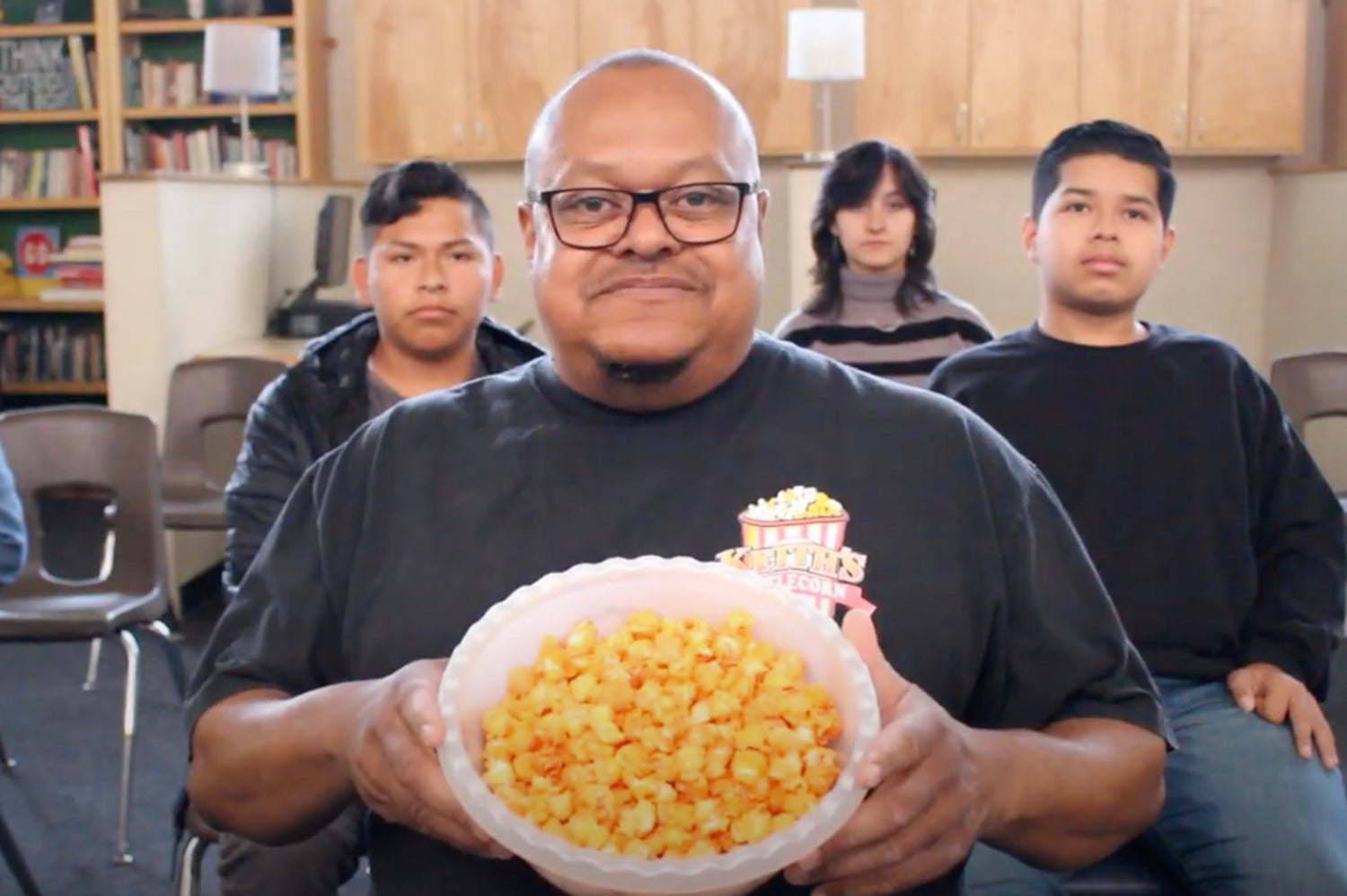 Keith Jackson, the founder of Keith's Kettle Corn, is shown in the image holding a large bowl of popcorn with a warm smile. He is wearing a black T-shirt with the Keith's Kettle Corn logo. Behind him, three young individuals sit attentively in a classroom-like setting, with bookshelves and cabinets in the background, suggesting an engaging and community-focused atmosphere.