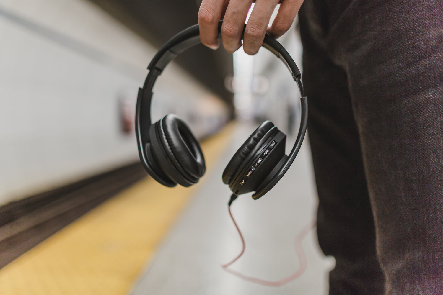 Close up of hands holding a pair of headphones, with the person standing on a subway platform while wearing black jeans